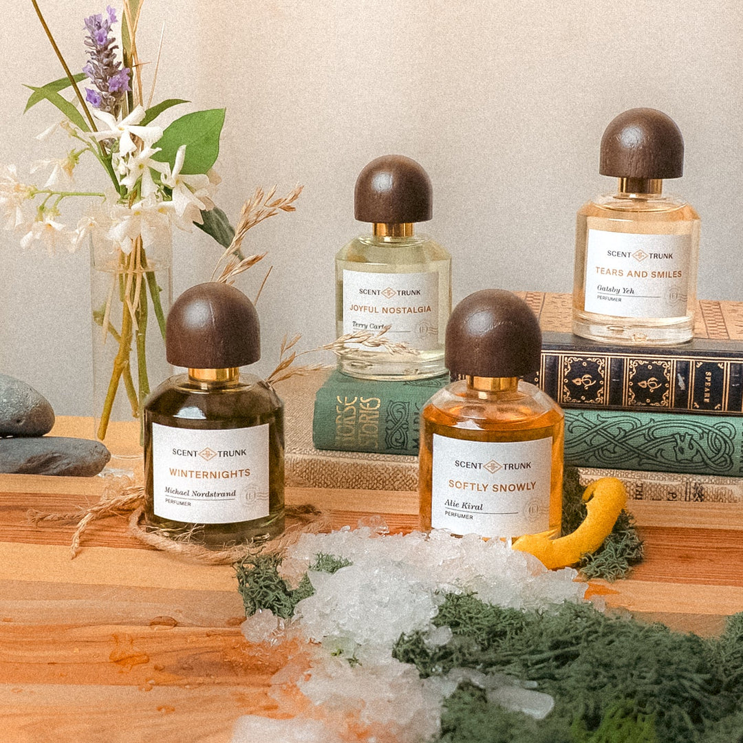 Three perfume bottles with dark wooden caps are arranged on a wooden surface with books, crystals, greenery, and flowers in a styled indoor setting.